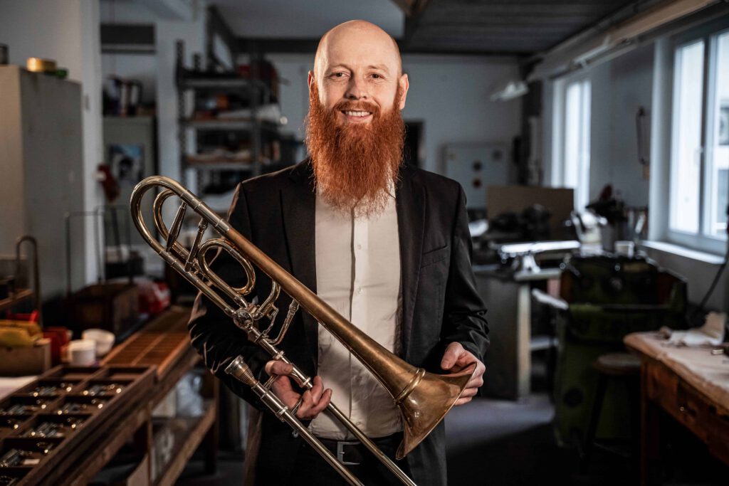 Hannes Hölzl Posaunist lächelnd im Kühnl Trombone Quartet mit Posaune in der Werkstatt der Blechblasinstrumentenfirma Kühnl & Hoyer